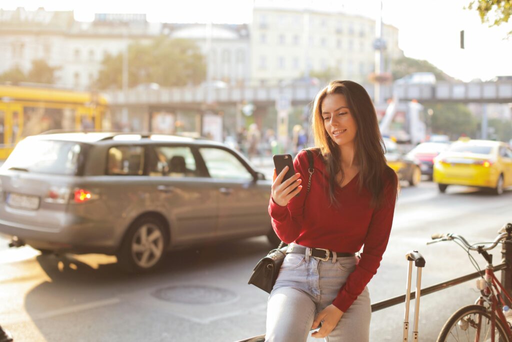 Smiling woman in a red sweater uses her phone on a sunny urban street. Cars and bicycles surround her.